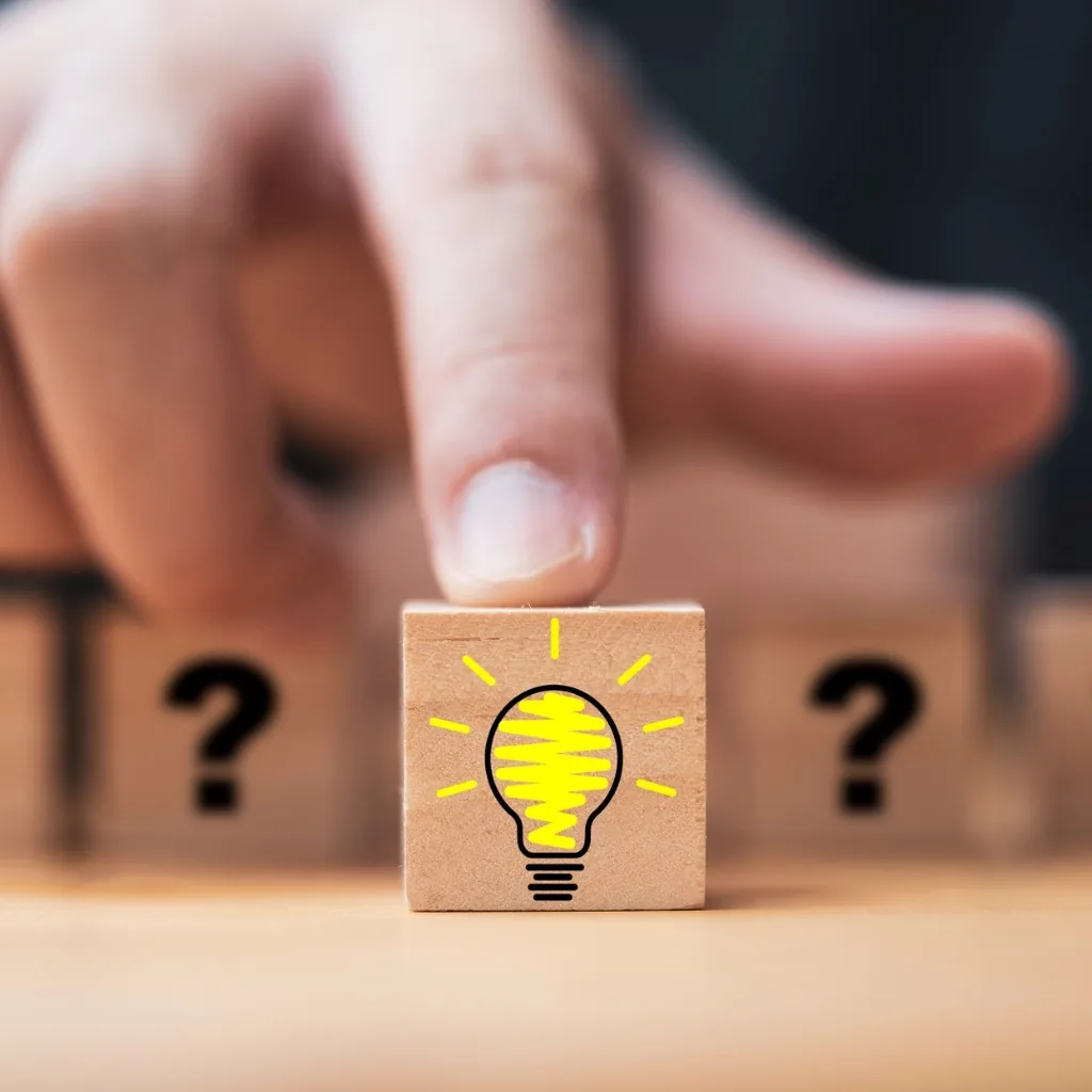 A hand placing a wooden block printed with a lightbulb among blocks printed with question marks